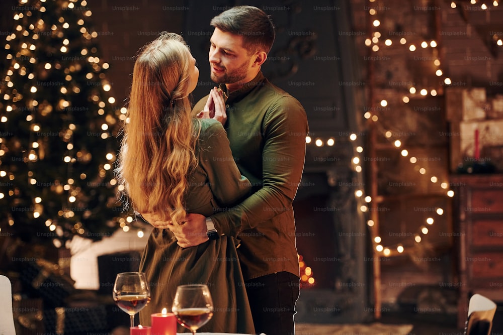 De pie en la habitación decorada con Navidad. Una joven y encantadora pareja tiene una cena romántica juntos en el interior.