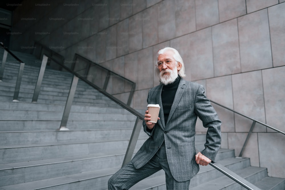 Un homme d’affaires âgé en tenue de soirée, avec des cheveux gris et une barbe est à l’extérieur et tient une tasse de boisson.