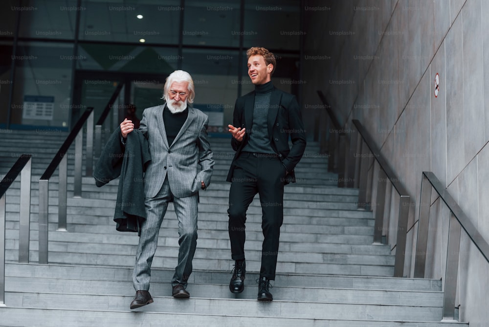 Walking on the stairs. Young guy with senior man in elegant clothes is outdoors together. Conception of business.