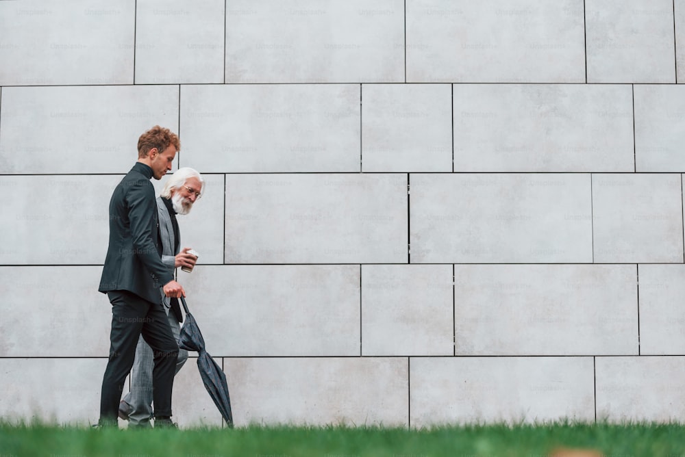 Walking on grass near wall. Young guy with senior man in elegant clothes is outdoors together. Conception of business.