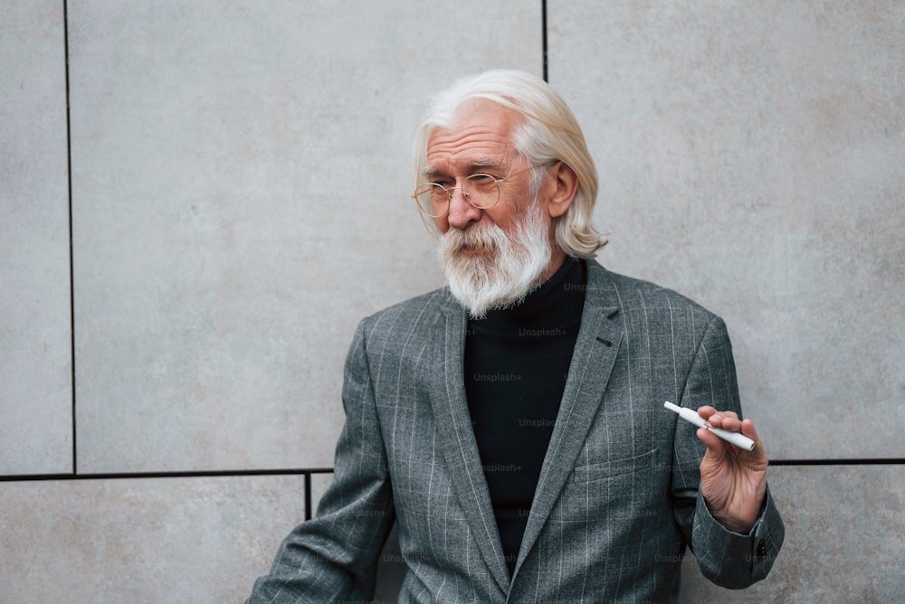 Senior businessman in formal clothes, with grey hair and beard is outdoors smokes electronic cigarette.