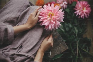 린넨 드레스를 입은 여자가 나무 소박한 벤치에 앉아 분홍색 달리아 꽃을 들고 있습니다. 시골의 느린 삶의 미학. 시골의 가을 시즌. 꽃집 가을 꽃다발 준비