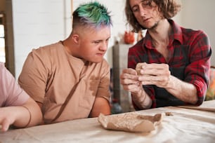 Do like this. Two men making ceramic pottery together. Craftsman in apron teaching young boy with down syndrome making plate from clay. Stock photo