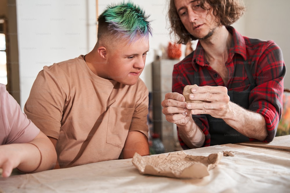 Hazlo así. Dos hombres haciendo cerámica juntos. Artesano en delantal enseñando a un niño con síndrome de Down a hacer un plato de arcilla. Foto de archivo