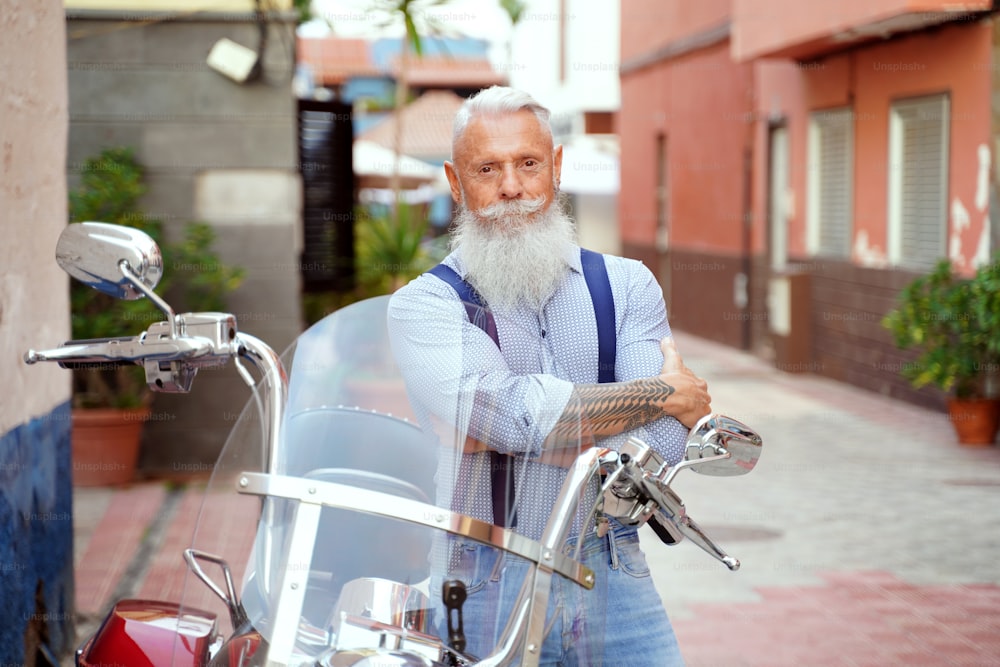 Hombre guapo con barba blanca posando cerca de una motocicleta en la calle de la ciudad, mirando a la cámara.