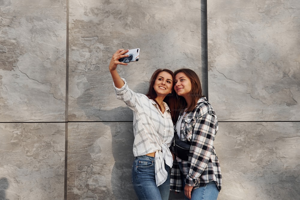 Fare selfie. Belle amiche allegre o coppia lesbica insieme vicino al muro all'aperto durante il giorno.