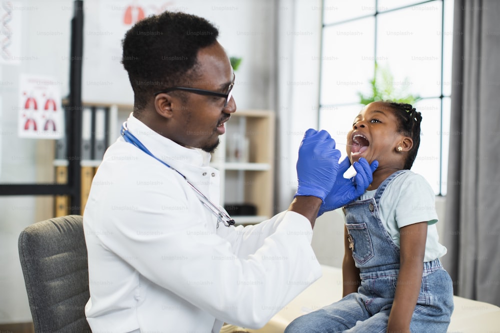 医務室のソファに座っている素敵な小さなアフリカ系アメリカ人の子供の女の子、ハンサムな黒人男性医師がへらで彼女の喉と口を調べている間。小児科のコンセプト
