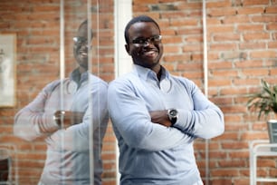 Confident black businessman standing with crossed arms at corporate office and looking at camera.