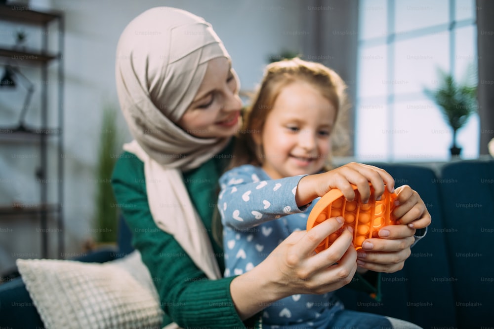 Orange antistress sensory toy fidget push pop it. Beautiful Muslim woman in hijab, sitting on sofa at home and playing with colorful pop it fidget toy with her little cute daughter. Focus on poppit