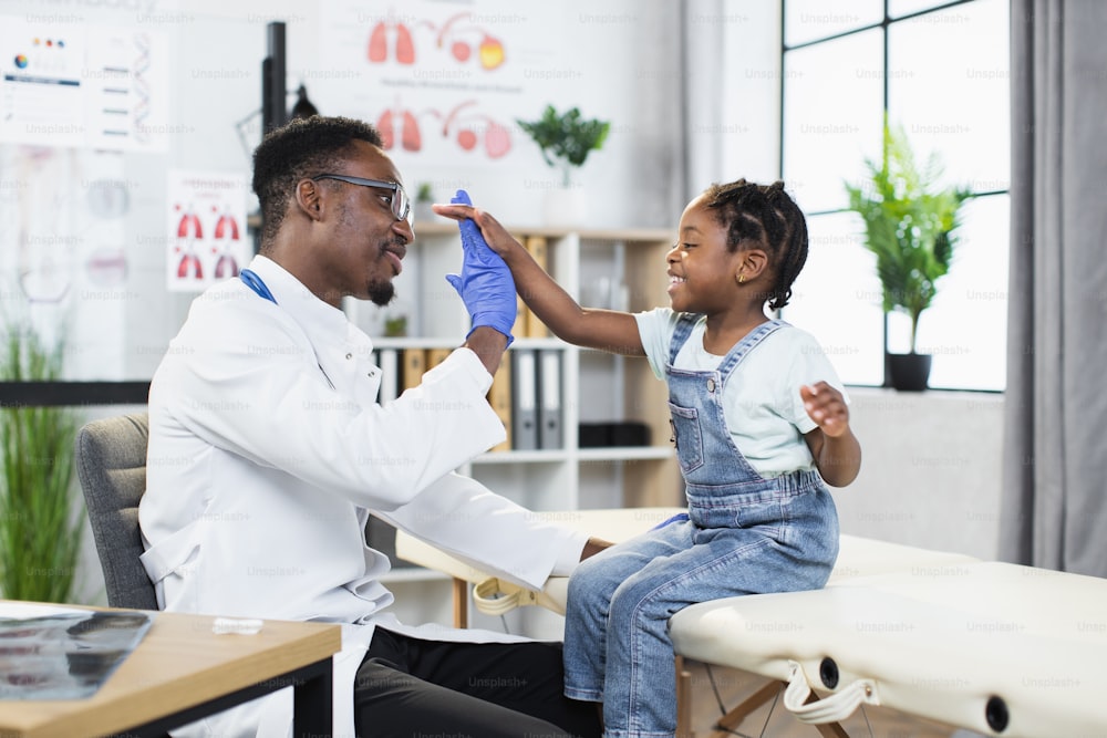Pretty girl giving high five to african male doctor after successful checkup at clinic. Competent pediatrician providing professional service to little patient.