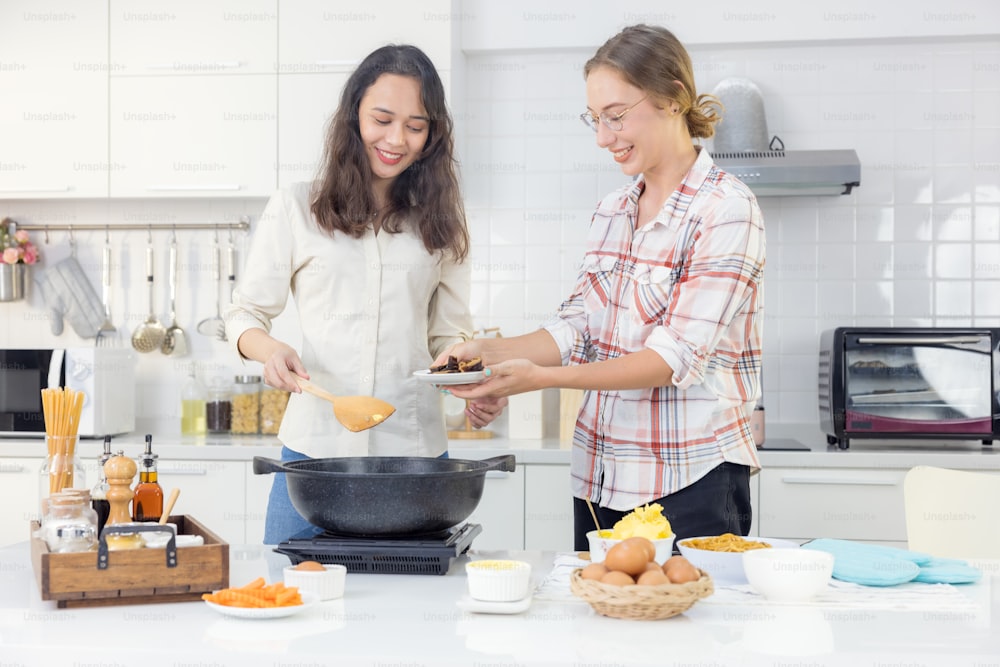 Una gioiosa giovane coppia lesbica in grembiule cucina insieme nella cucina di casa dietro un tavolo di legno con una padella e una spatola.