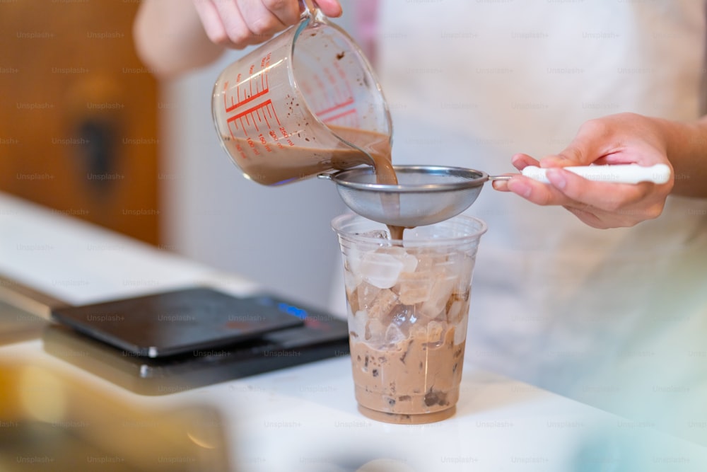 Close up hand of Asian man coffee shop part time employee making iced cocoa with froth milk at bar counter. Male barista preparation cold drink for customer at cafe. Small business owner and part time job concept