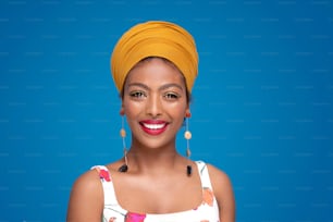 Hermosa chica étnica afroamericana feliz con turbante en la cabeza sonriendo a la cámara, posando sobre fondo de estudio azul pastel. Cultura. Emociones de personas reales. Moda. Retrato colorido.