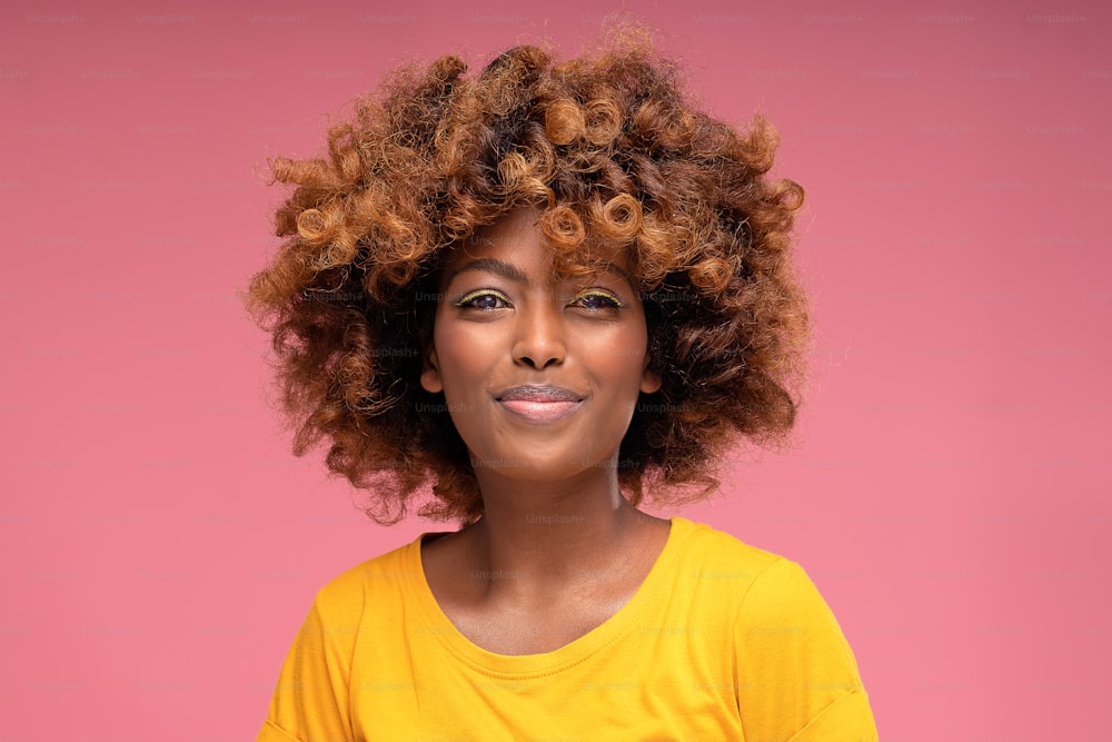 Heureuse belle fille afro avec un maquillage d’été glamour et des cheveux bouclés souriant à la caméra sur fond de studio pastel rose. Photo colorée. De vraies émotions de personnes.
