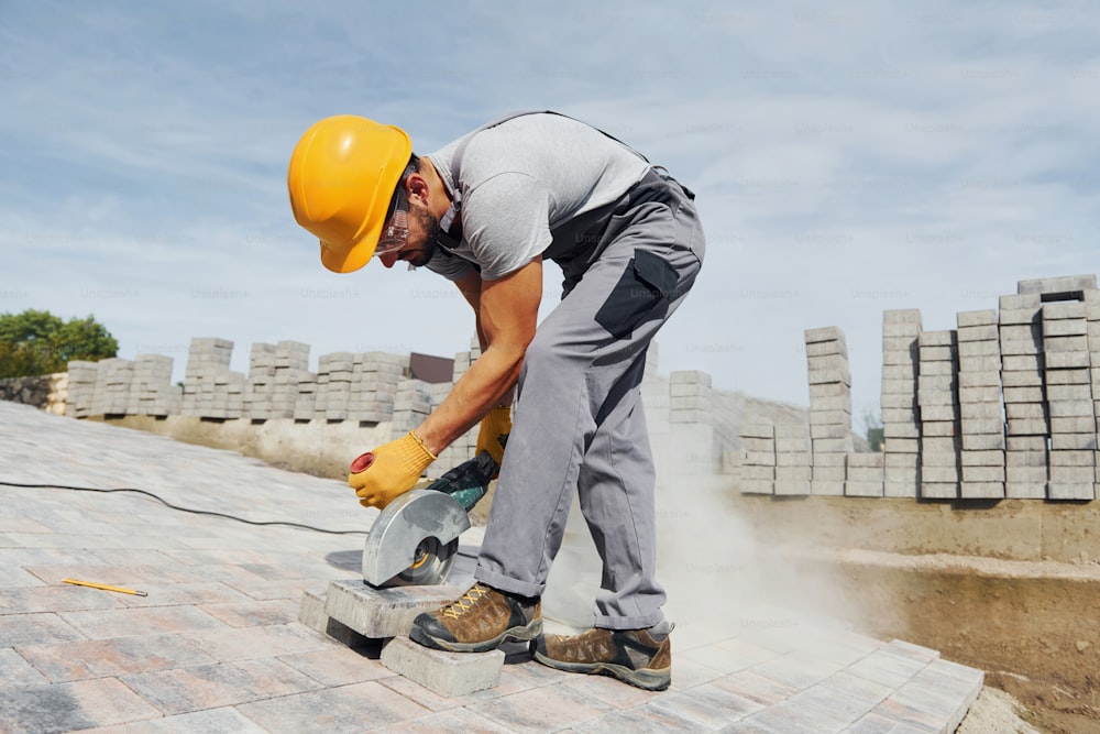 Uses circular saw. Male worker in yellow colored uniform have job with pavement.