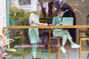 Asian woman coffee shop waitress barista serving soft drink and bakery to customer on bar counter at cafe. Businesswoman or freelancer sitting by the window working on laptop computer at restaurant. Small business and part time job concept