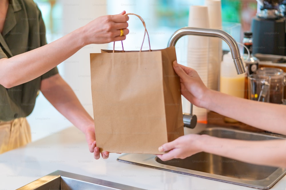Young Asian woman coffee shop employee barista working at cafe. Smiling female waitress or cashier preparing takeaway order in paper bag to customer. Small business owner and part time job working concept