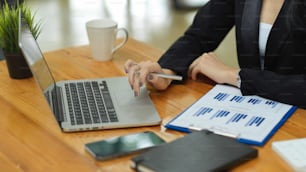 Mujer de negocios analizando las finanzas de la empresa a través de una computadora portátil con informe financiero, teléfono inteligente, libro de planificación y taza de café en el escritorio de madera en la oficina