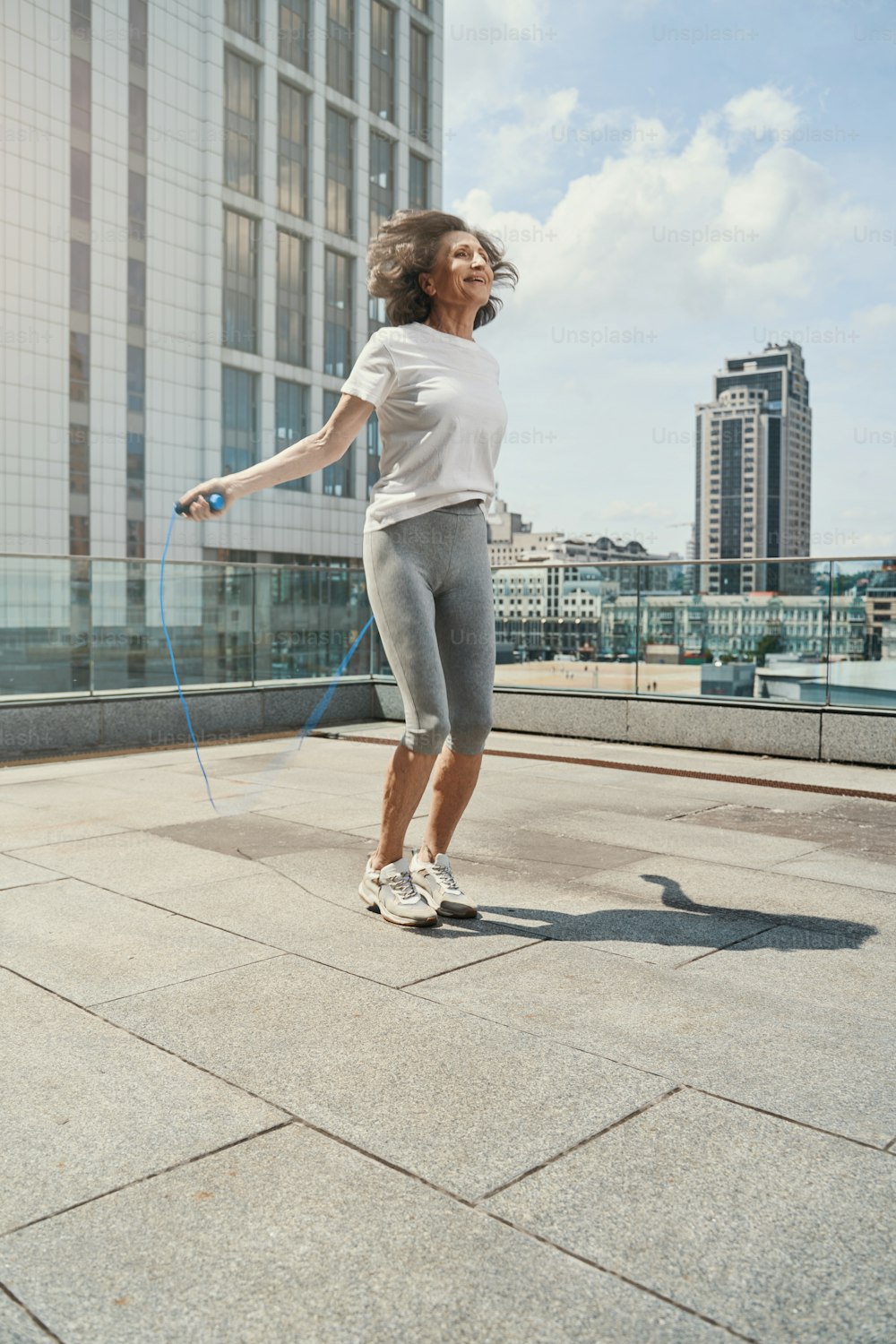 Fröhliche sportliche Dame verbringt aktive Zeit mit Springseil auf offener Terrasse über der Stadt