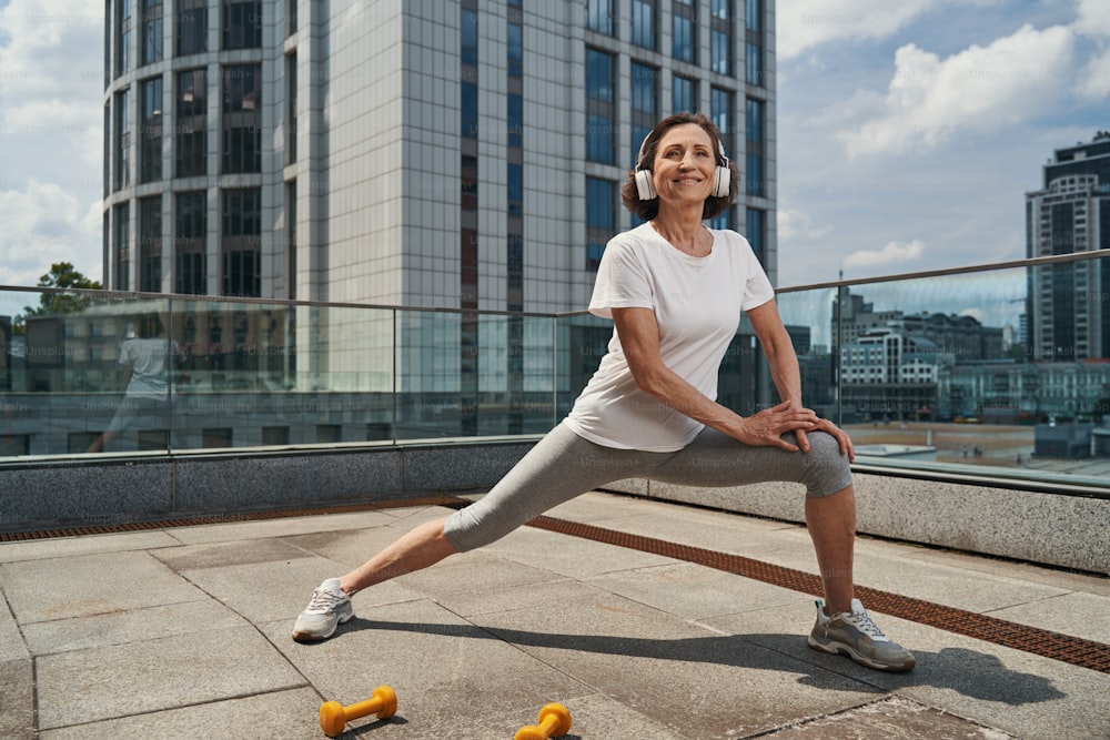 Smiling active senior woman is doing lunges while listening to music with headphones in open air