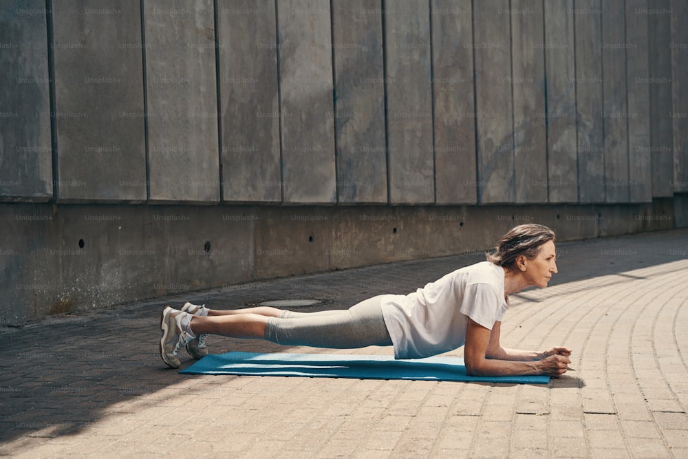 Una mujer madura relajada está ejercitando el núcleo en la colchoneta mientras entrena al sol cerca de una pared de hormigón