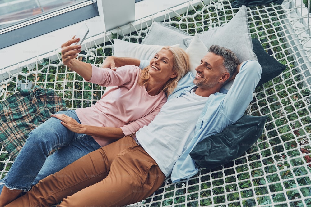 Vista dall'alto di bella coppia matura che fa selfie con lo smartphone e sorride mentre si rilassa insieme in una grande amaca a casa