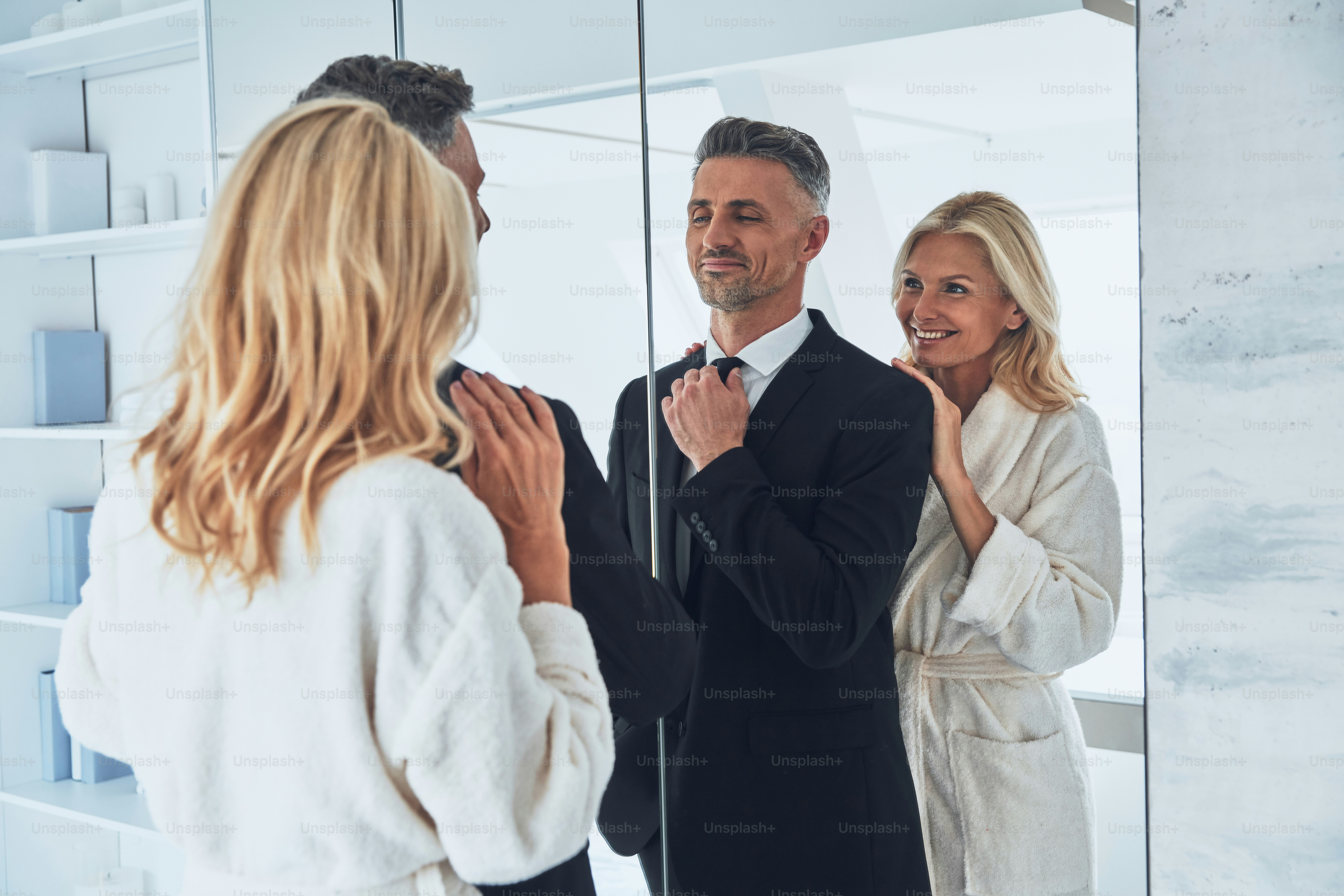 Beautiful mature woman adjusting suit of her husband while both standing in front of the mirror at home