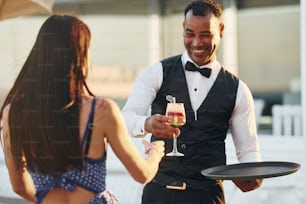 Order for woman. Black waiter in formal clothes is at his work outdoors at sunny daytime.