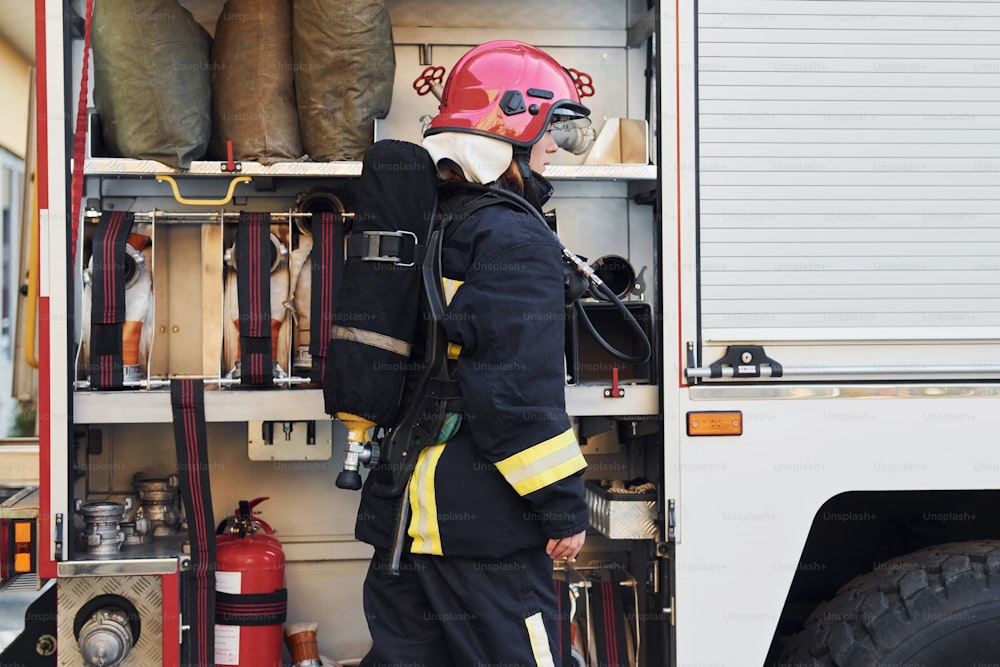 Takes equipment. Female firefighter in protective uniform standing near truck.