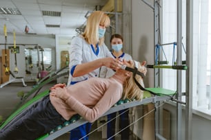 Female doctor rehabilitologist working with female patient on the special rehabilitation device at rehabilitation center gym. Doctor and nurse wearing protective face masks.