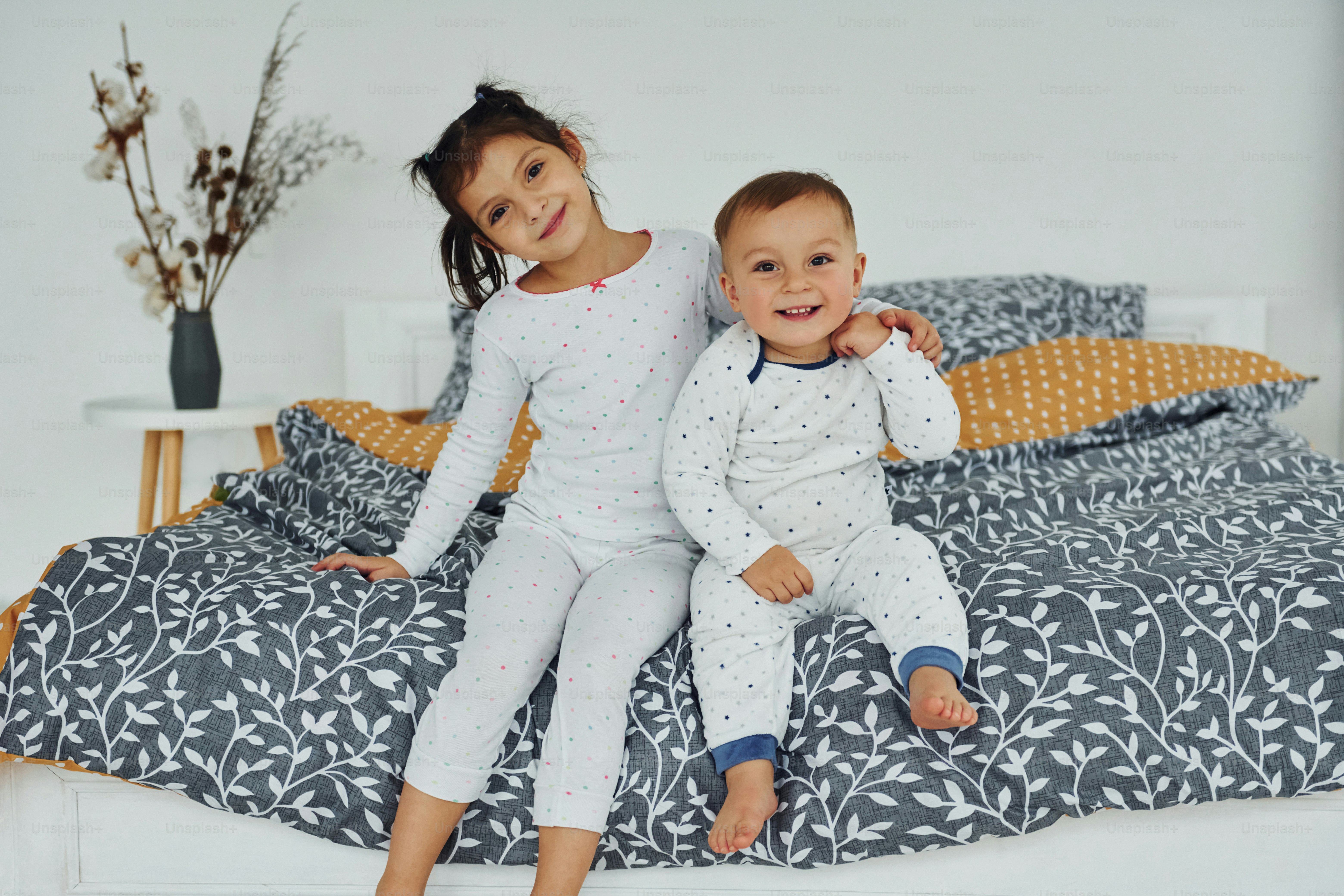 Little sister and brother sits together on the bed. Interior and design of beautiful modern bedroom at daytime.