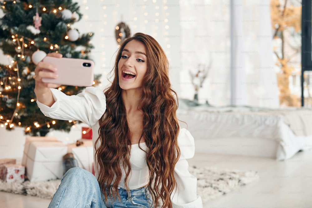 Happy young woman sits indoors near christmas tree and celebrates New year.
