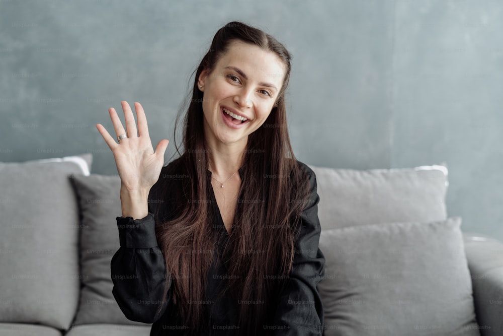 Happy young woman blogger looking at camera and waving hello while recording video for social media or her vlog, greeting followers while sitting on comfortable sofa at home