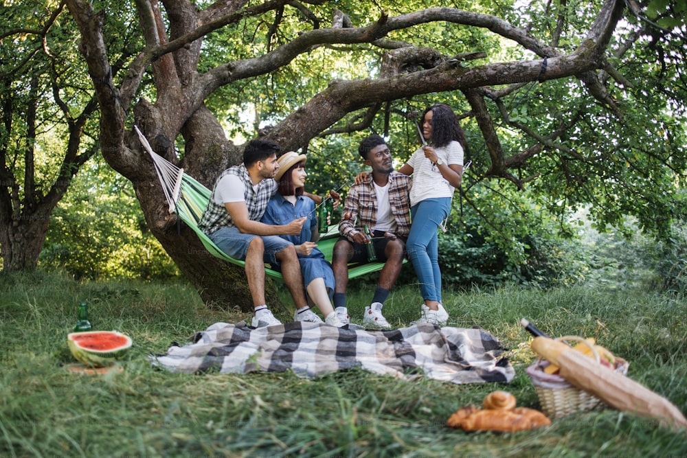 Group of four multicultural friends eating food and drinking beer during leisure time on nature. Young people having picnic at summer garden.