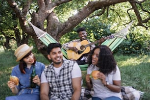 African american guy sitting in hammock and playing guitar while his three multicultural friends relaxing on grass with drinks and snacks. Concept of people, leisure and nature.
