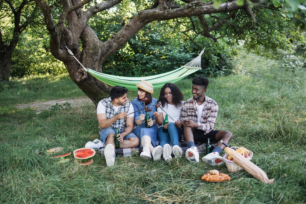 Group of happy international people in summer clothes having picnic at garden with cold beer and grilled sausages. Concept of friendship, leisure and togetherness.