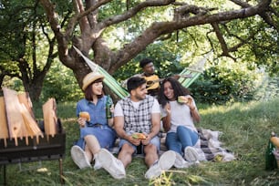 Three young people sitting on plaid at summer garden eating tasty burgers and drinking beer. Blur background of african guy playing guitar in hammock. Picnic of diverse friends.