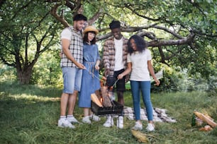 소시지를 굽기 위해 바베큐 주위에 모인 4 명의 다문화 친구들. 여름 옷을 입은 젊은이들이 야외에서 불에 음식을 요리하고 있습니다.