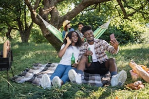 African american couple sitting at green garden and taking selfie on modern smartphone while their multiracial friends talking in hammock on background. Summer picnic outdoors.