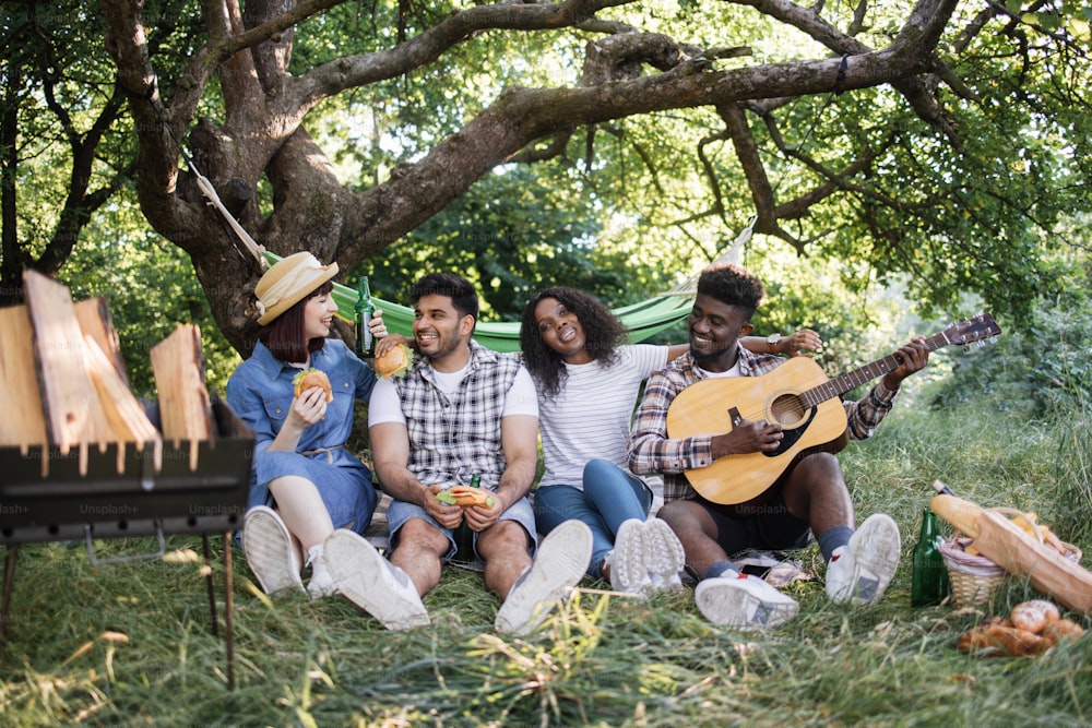 Two multiracial couple singing and playing guitar while resting outdoors. Happy friends having picnic with food, drinks and entertainment.