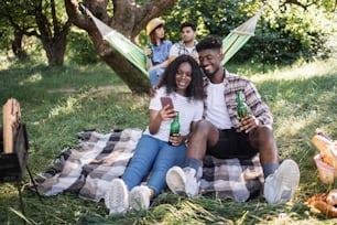 사랑스러운 흑인 커플은 정원에서 포옹을 하고 모바일 화면을 보고 다양한 친구들이 해먹에서 이야기하고 술을 마시고 있습니다. 자연에서 피크닉을 즐기는 네 친구.