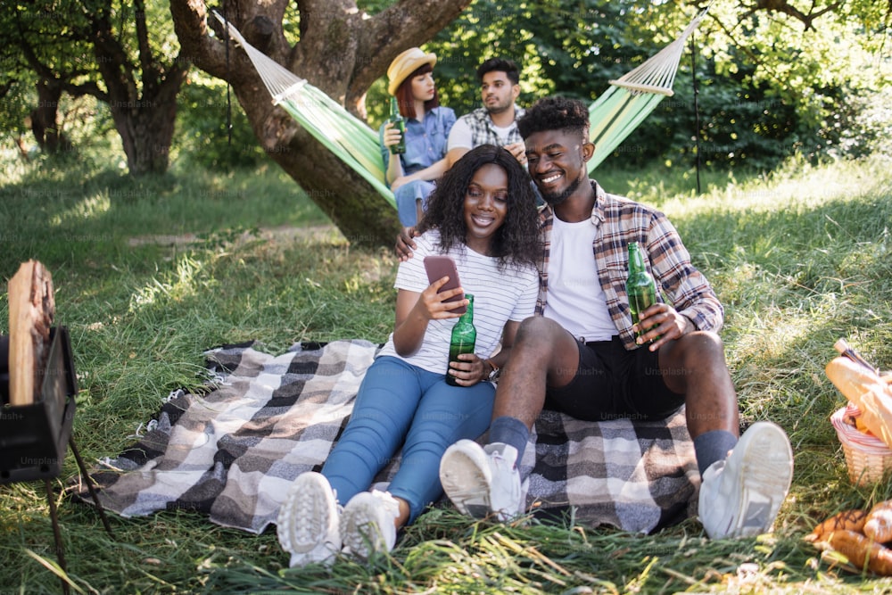 Lovely black couple sitting in hugs at garden and looking on mobile screen while their diverse friends talking and drinking in hammock. Four friends having picnic on nature.