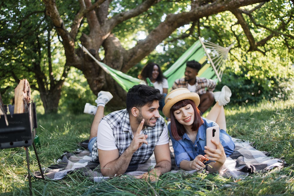 Cheerful couple taking selfie on smartphone during picnic outdoors. Blur background of african man and woman sitting in hammock and drinking beer. Summertime and friendship concept.