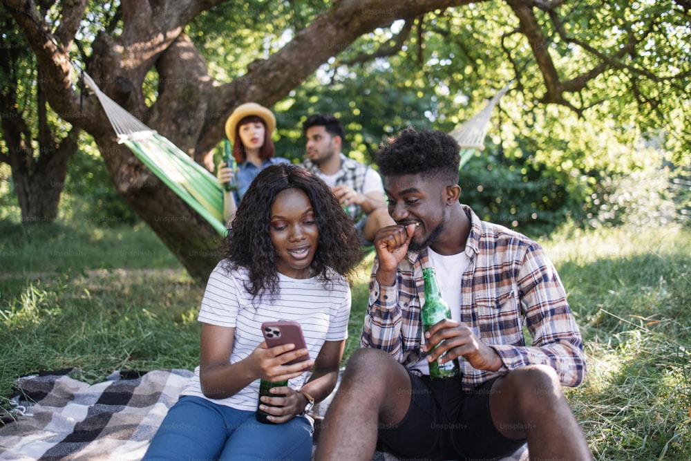 아프리카계 미국인 커플은 여름 정원에서 격자무늬에 함께 앉아 현대 스마트폰을 사용하고 있다. 다인종 친구들이 배경에서 해먹에서 수다를 떨고 맥주를 마시고 있다.