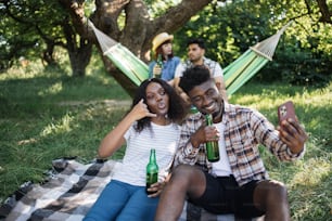재미있는 흑인 남자와 여름 공원에서 휴식을 취하고 모바일에서 셀카를 찍는 여자. 맥주를 손에 들고 해먹에 앉아 있는 두 명의 다양한 친구의 배경을 흐리게 합니다.