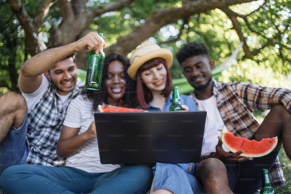 Group of four multi ethnic people using wireless laptop for video conversation outdoors. Happy friends enjoying picnic time with modern technology, food and drinks.