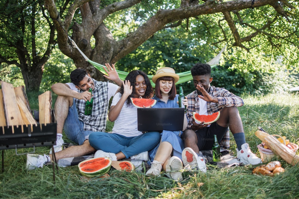 Cuatro amigos multirraciales sonriendo y saludando durante una videollamada en una computadora portátil moderna mientras se relajan en la naturaleza. Jóvenes con alcohol y comida haciendo un picnic en el jardín.