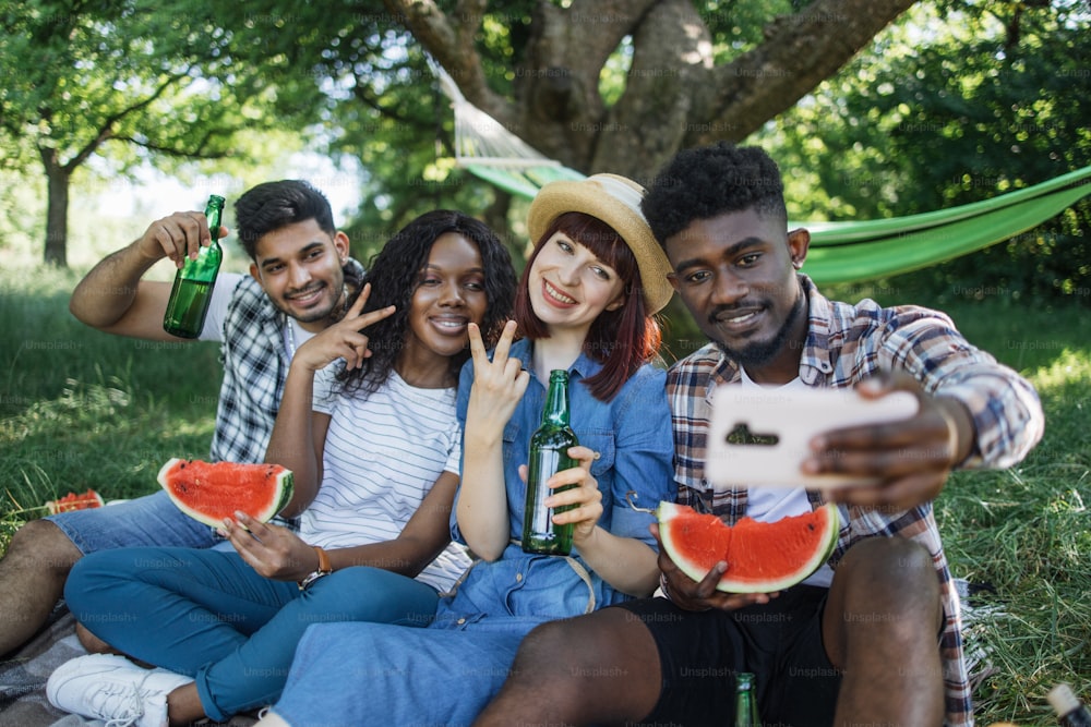 Cheerful group of four diverse friends sitting at green garden with watermelon and beer in hands and taking selfie on modern smartphone, Concept of people, technology and leisure time.