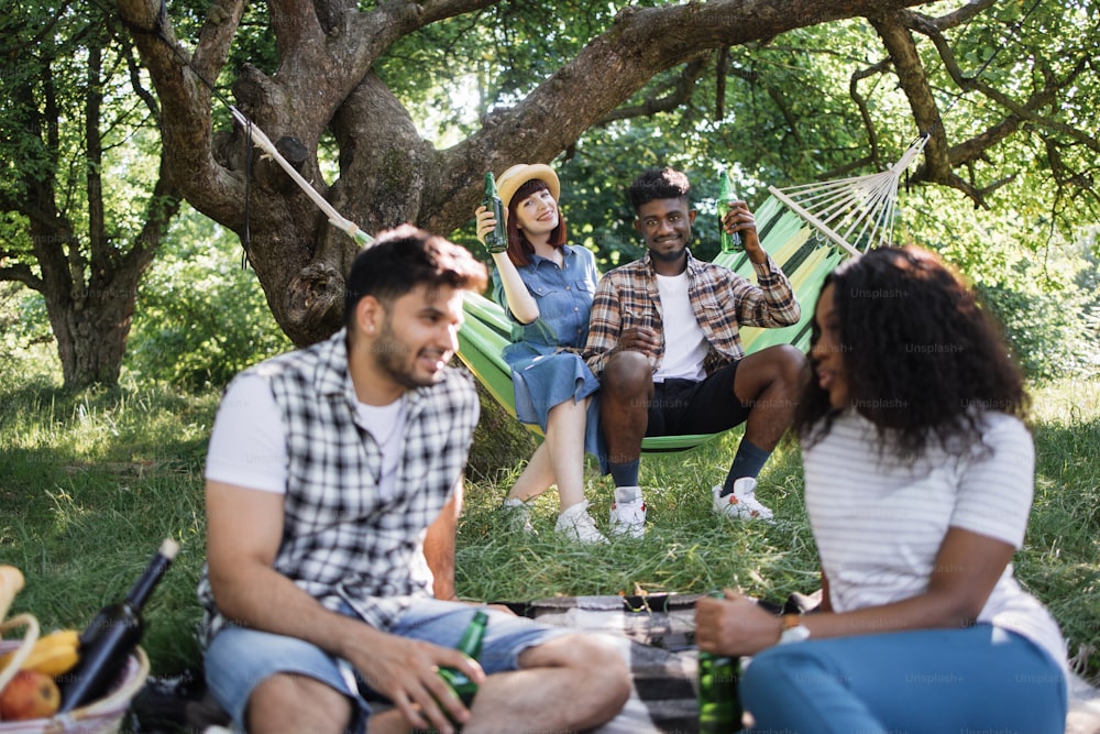 Relaxed multiracial people in casual clothes spending free time on fresh air with tasty food and alcohol. One couple sitting on grass, another resting in hammock. Summertime concept.