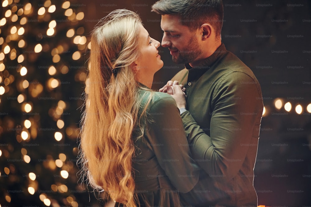 De pie en la habitación decorada con Navidad. Una joven y encantadora pareja tiene una cena romántica juntos en el interior.
